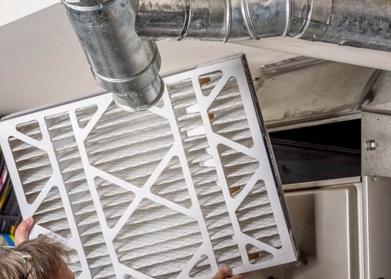 A person is inserting a new furnace filter. The ducting leading to the furnace is visible.