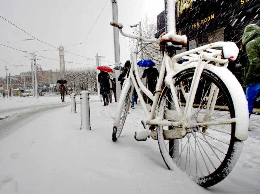 Photo of a bicycle parked next to a sidewalk. The sky is gray and the sidewalk, street and bike are covered in snow.