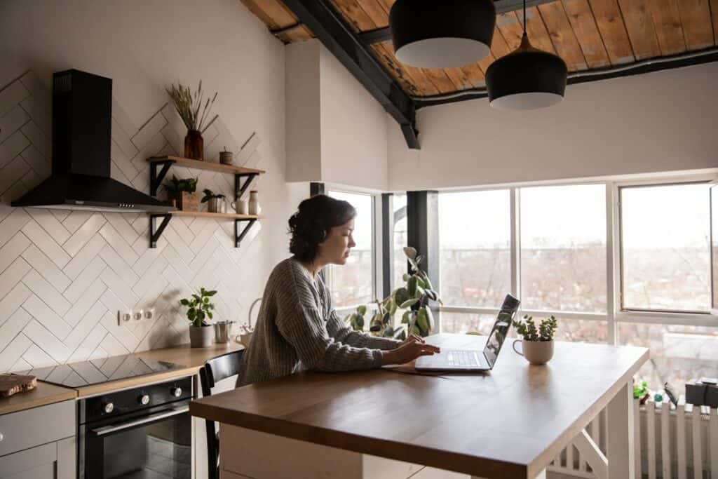Photo of a young woman standing in a modern kitchen using her laptop on the counter, researching reasons why her air conditioner is turning on and off.