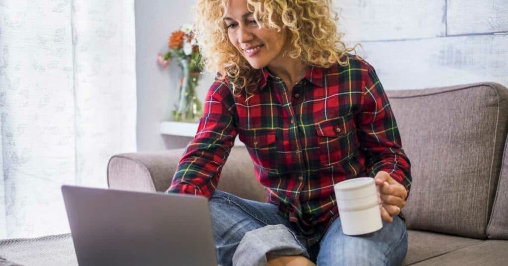 a woman sits on a sofa, holding a coffee cup and looking at a laptop