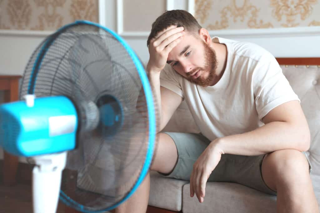 Young bearded man using electric fan at home, sitting on couch cooling off during hot weather, suffering from heat, high temperature wonderingwhy is my air conditioner not blowing cold air