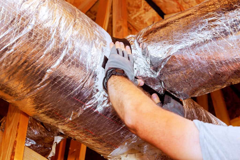 Man repairs insulated ductwork in a residential attic during daylight hours